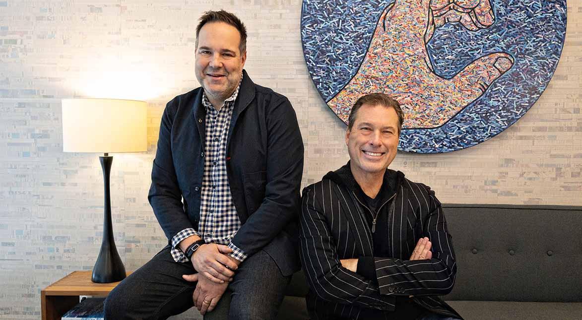 Two men smiling in a modern room; one is sitting on a couch with arms crossed, the other is sitting on the armrest. A lamp is on the left and a large textured artwork of a hand is on the wall behind them.